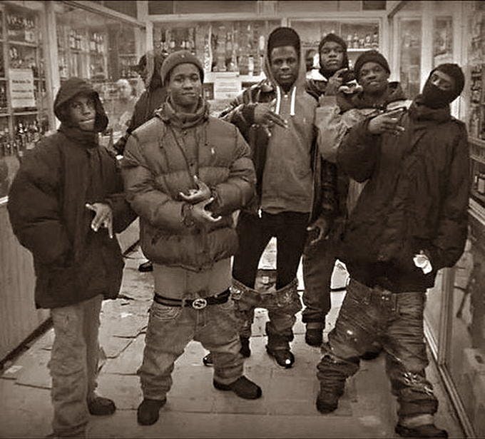 Grayscale photo of six Black men in a shop making gang signs.