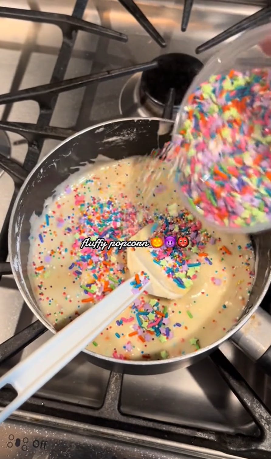 Fluffy popcorn being cooked on the stove top, the baker is pouring rainbow sprinkles into the pan.