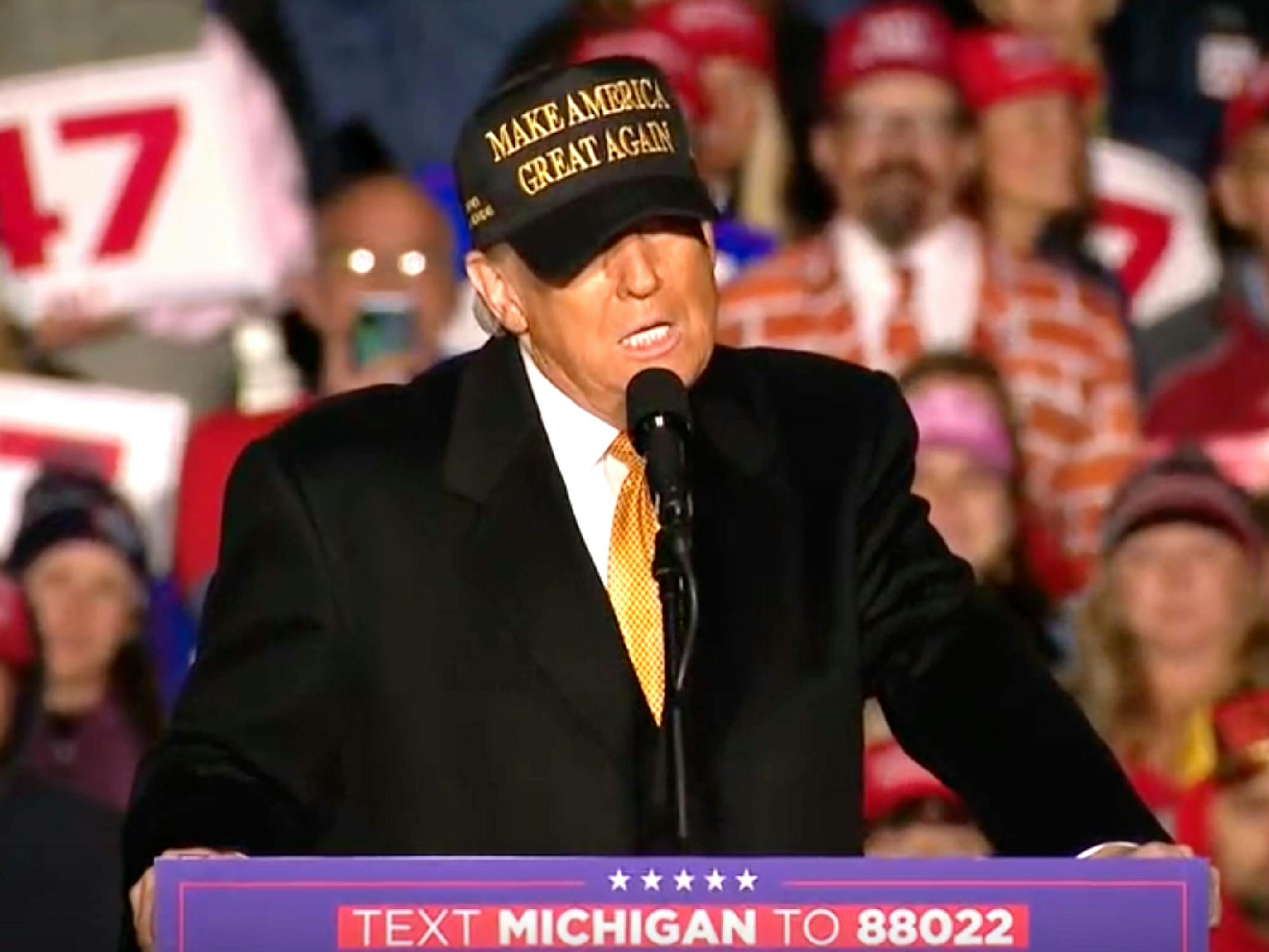 Donald Trump speaking in front of a Michiigan crowd. He stands at lectern and wears a new yellow and black 'Make America Great AGain' hat.