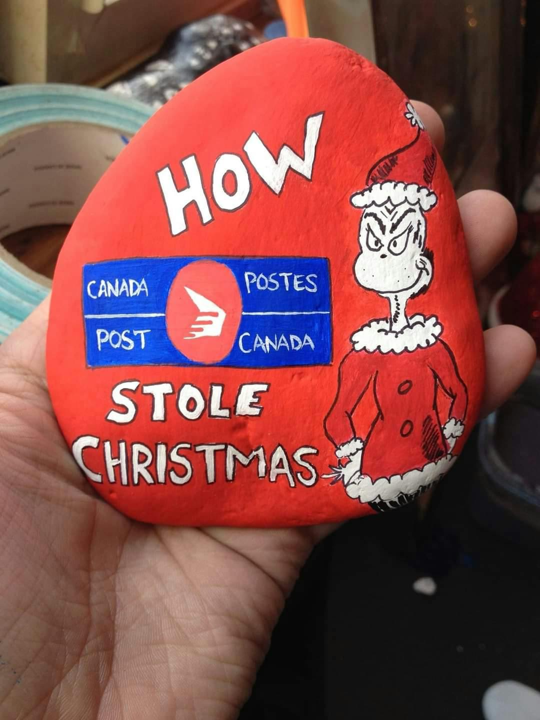 A painted rock in the Grinch's book art style, a drawing of the Grinch and Canada Post's logo. Text reads, 'How Canada Post stole Christmas.'