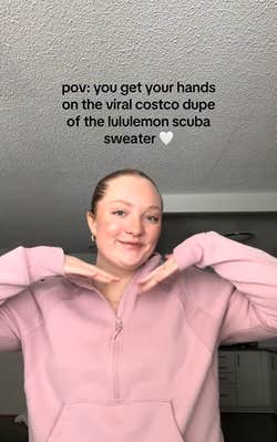 A blonde woman in a pink dupe Scuba hoodie posing for her camera. Text overlay reads, "POV: you get your hands on the viral costco dupe of the lululemon scuba sweater (heart emoji)"