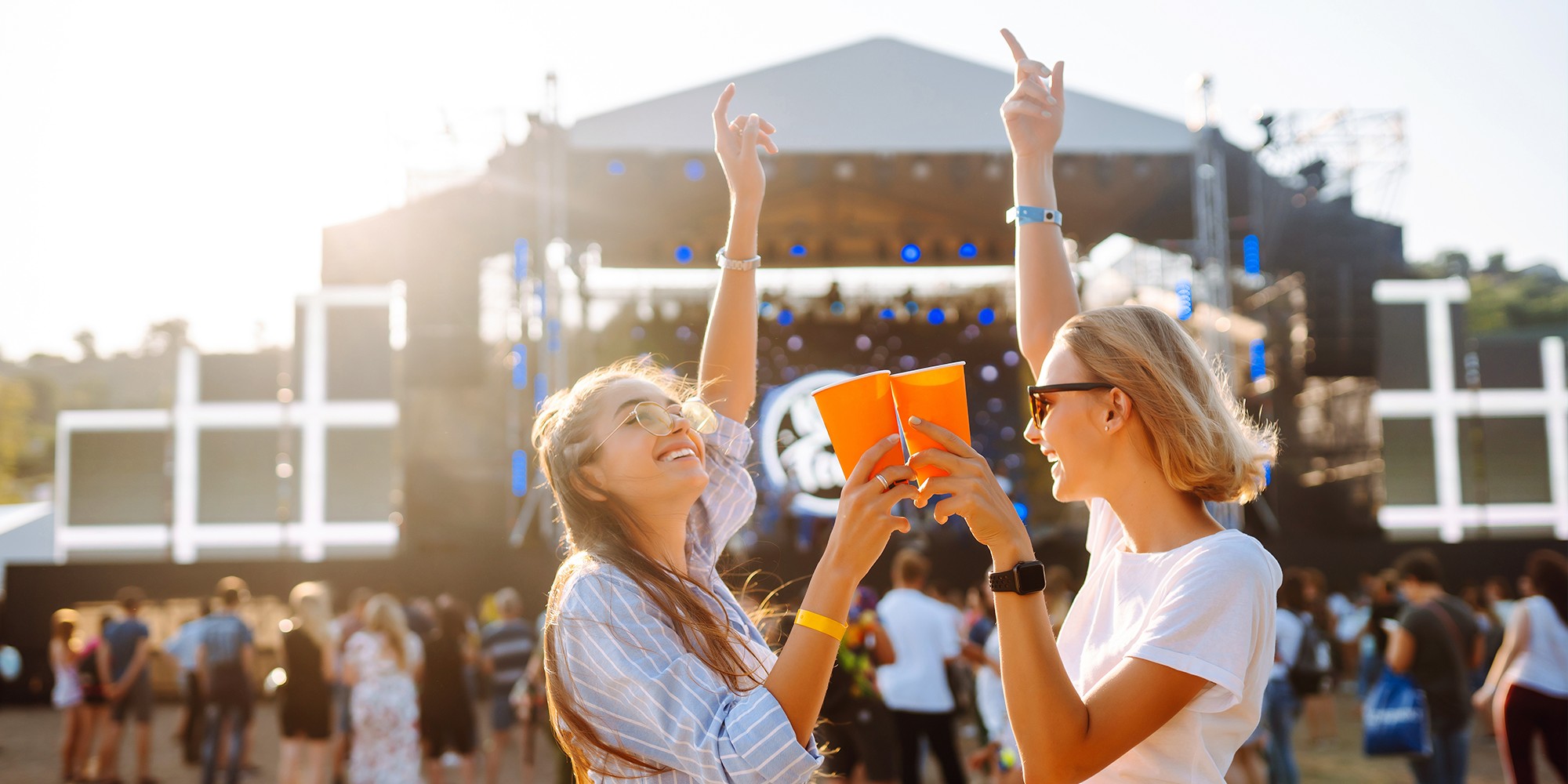 Two women hold drinks and point up to the sky at an outdoor music festival.