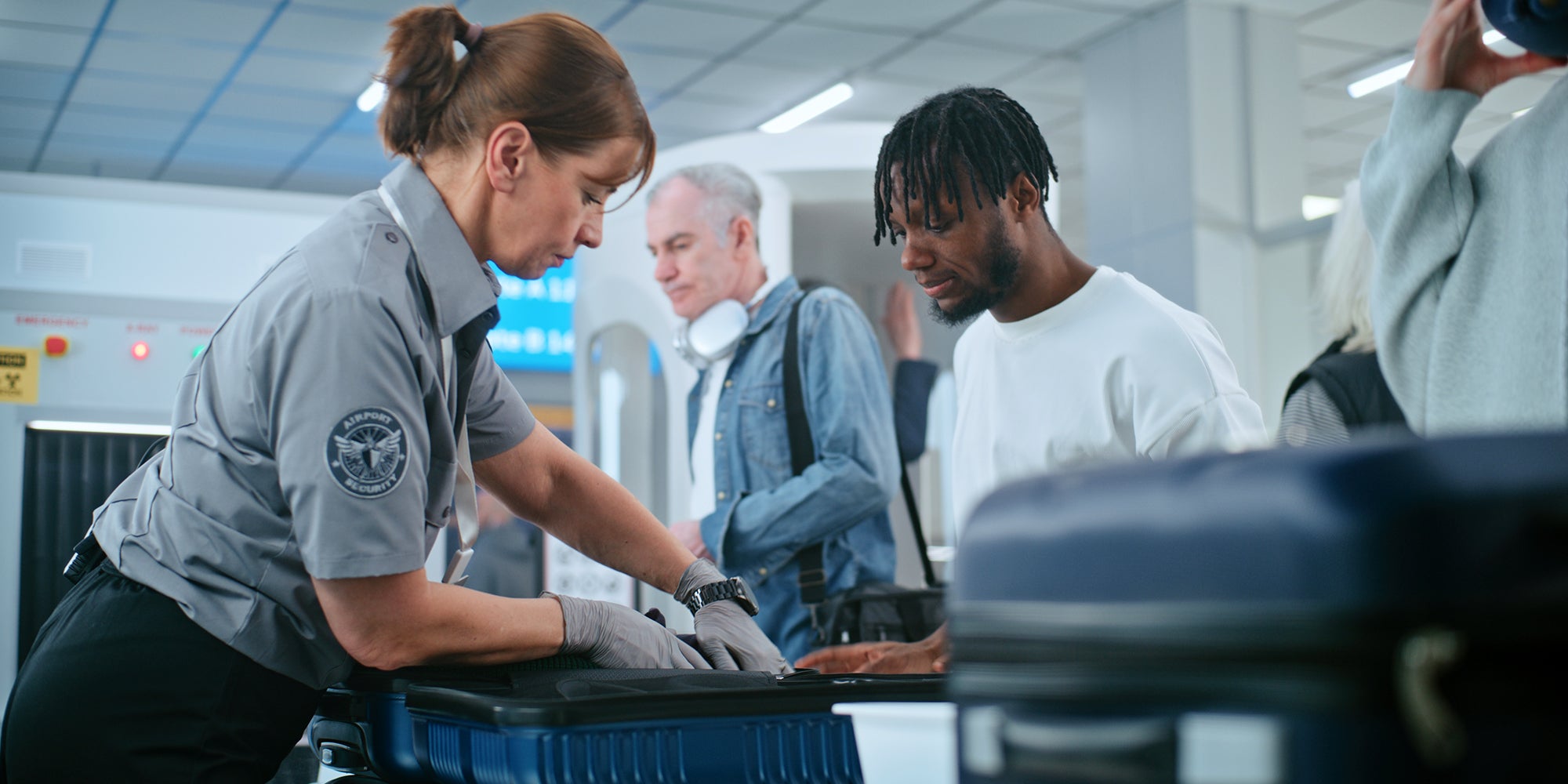 Security Checkpoint in Airport Terminal: Female TSA Worker Inspecting Baggage of Passenger before Boarding Flight, Finding and Confiscates Liquid. Queue of Diverse People During Screening Procedures.