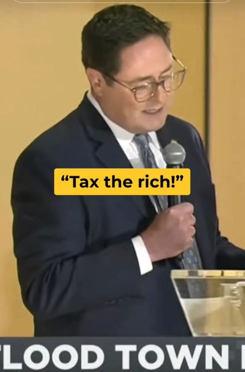 Rep. Mike Flood standing on stage with a mic and grimacing as the crowd chants 'Tax the rich!'