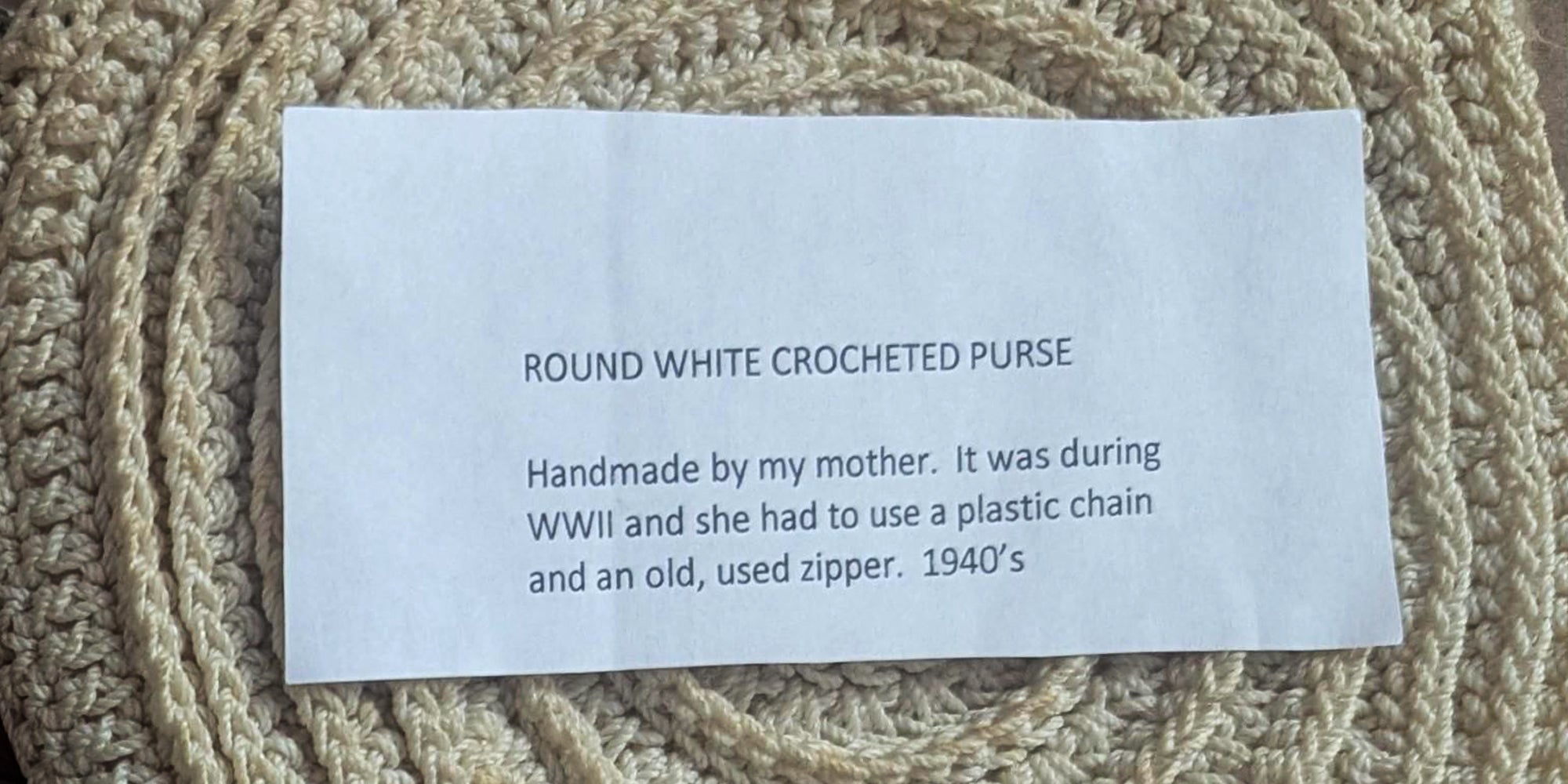 Crocheted white purse. Printed note sitting atop it reads, 'ROUND WHITE CROCHETED PURSE: Handmade by my mother. It was during WWII and she had to use a plastic chain and an old, user zipper. 1940's.'