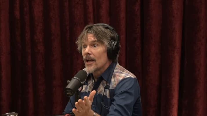 Ethan Hawke sitting in front of a red curtain and gesturing with his hands while he speaks into a microphone.