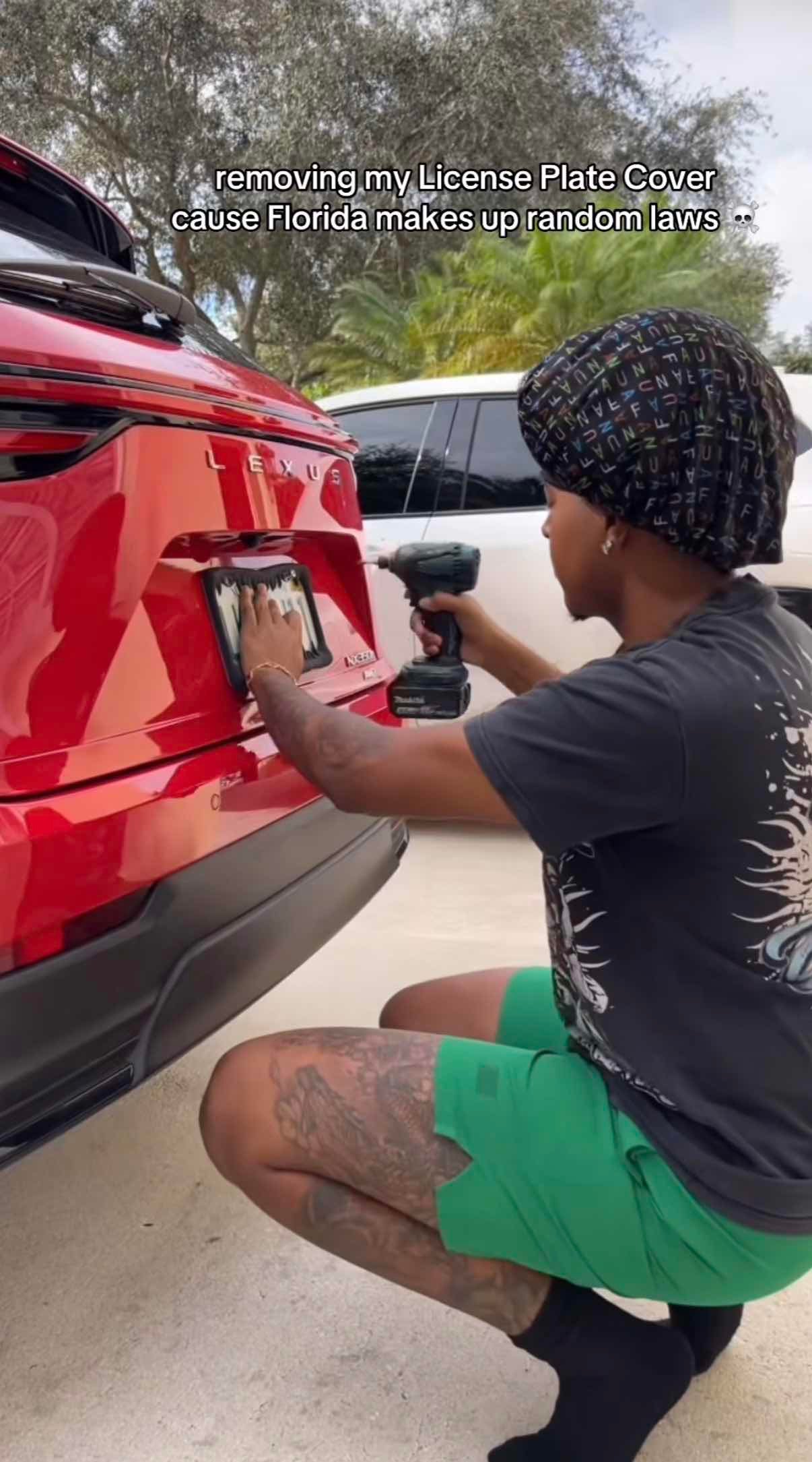 A man crouched down behind his car, carefully removing the license plate frame with a drill.