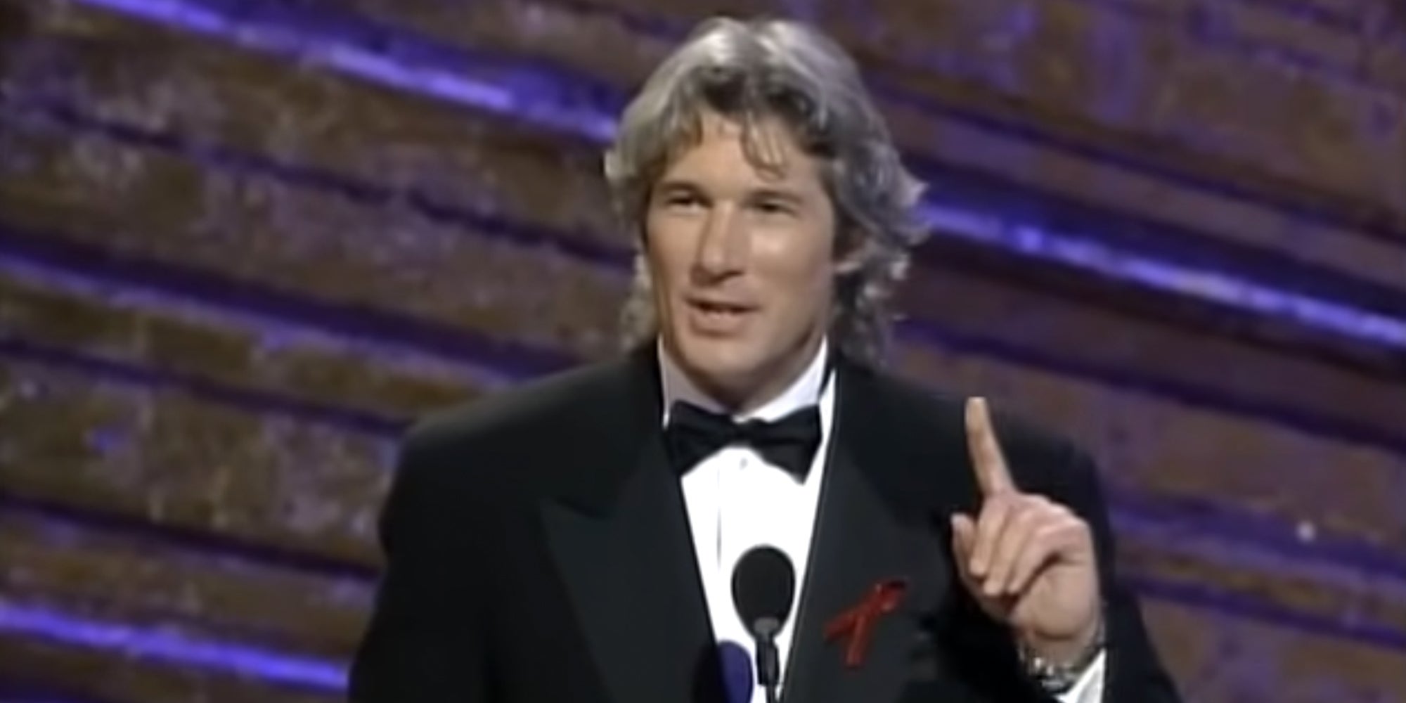Actor Richard Gere wearing a black tuxedo and speaking into the microphone at the 1993 Oscar Awards.