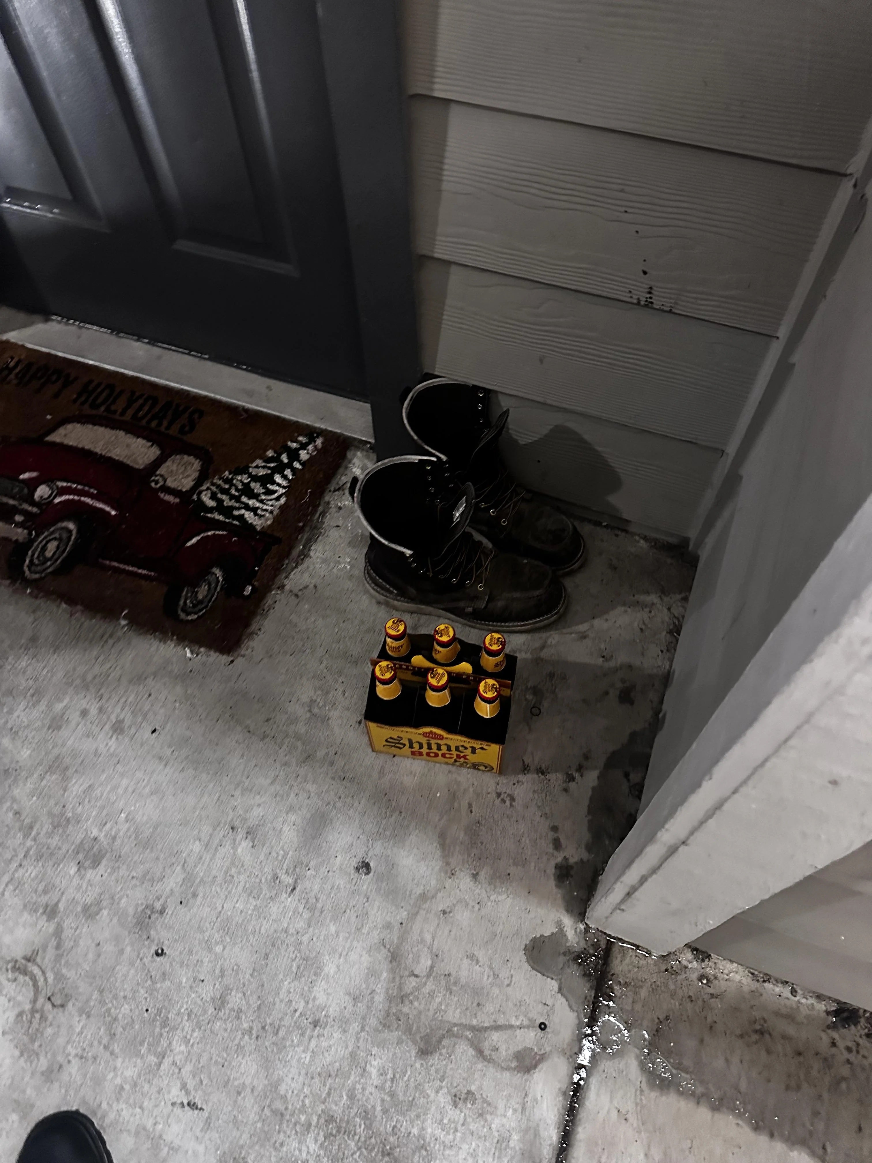 Photo of a six-pack of beer left by a pair of boots next to an apartment front door.