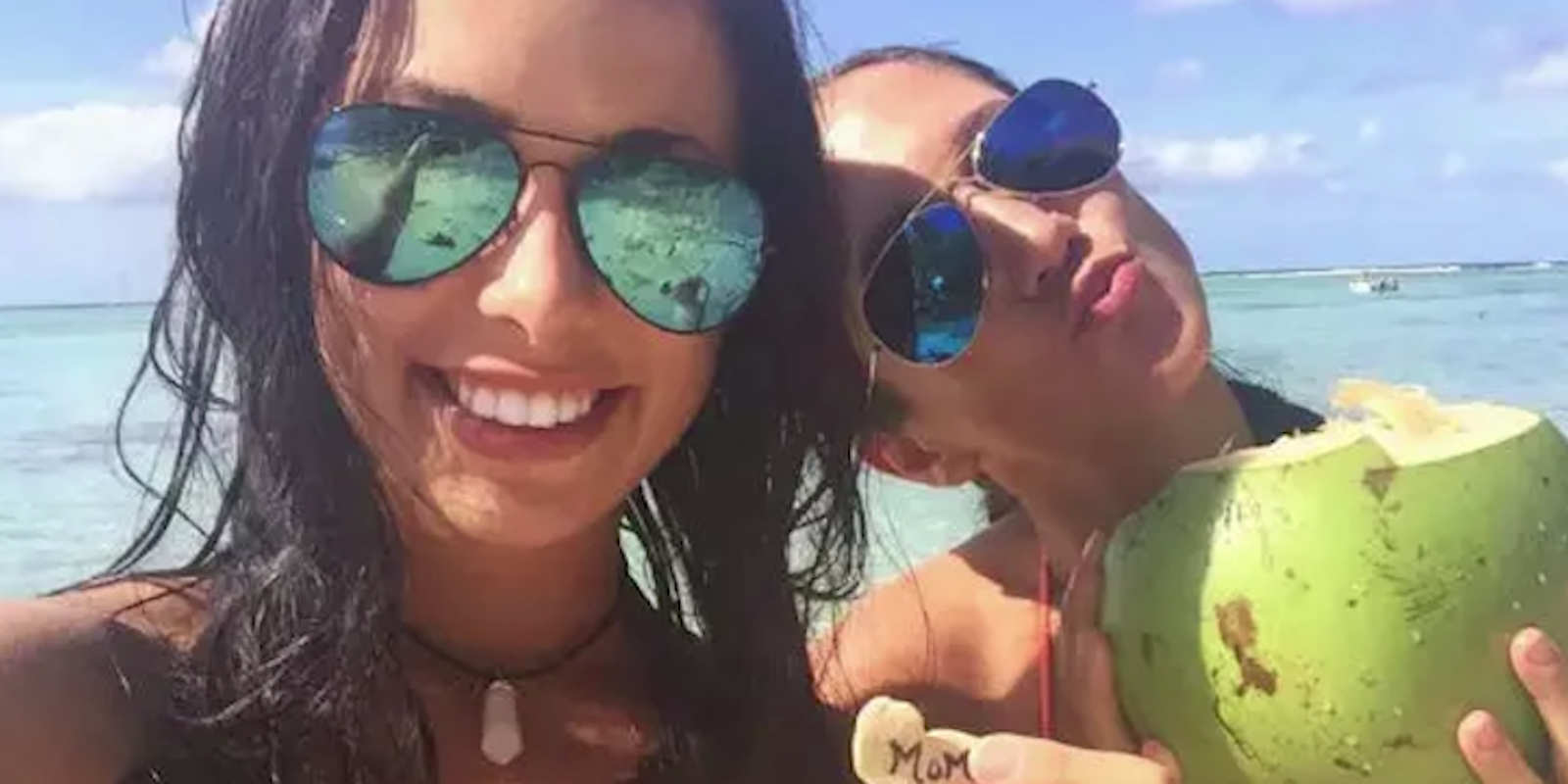 Two girls smile for a selfie in sunglasses, one holds a drink in a coconut