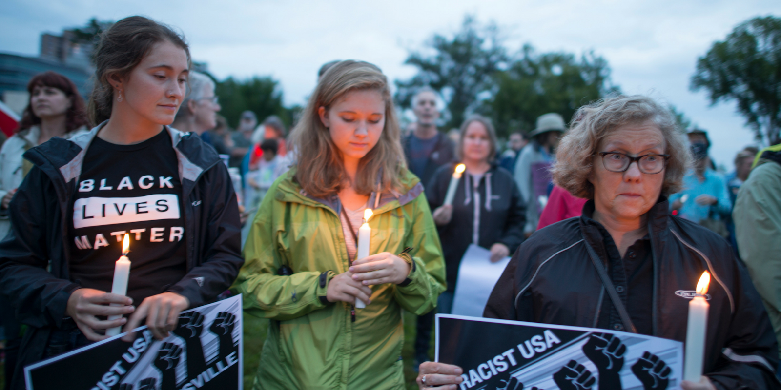 Three women stand with lit candles and anti-racist signs at a vigil for Charlottesville victims.
